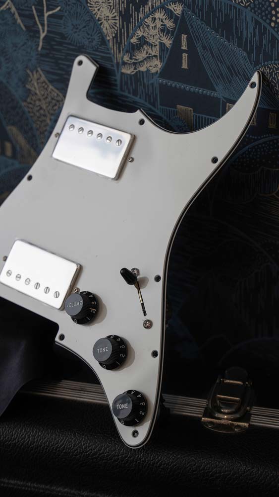 a parchment white loaded pickguard for HH Stratocaster is placed on top of a guitar case. The pickguard is rested against a wall with patterned blue and gold wall paper.